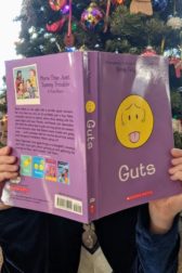 Child holding open a book to cover their face. The book is Guts by Raina Telgemeier