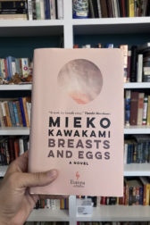 The pale pink cover of "Breasts and Eggs" held up in front of a large bookcase