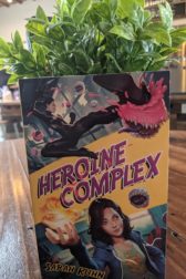 The book Heroine Complex by Sarah Kuhn sitting on a wooden table top with a leafy plant behind it.