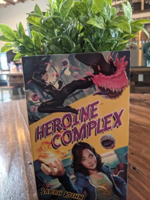 The book Heroine Complex by Sarah Kuhn sitting on a wooden table top with a leafy plant behind it.
