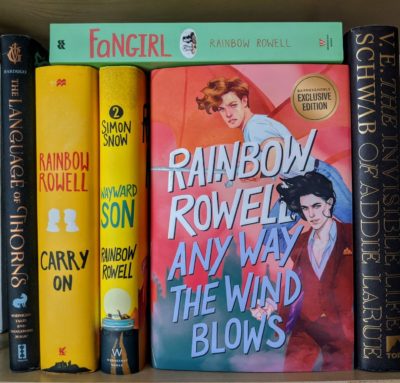 Several books on a bookshelf with "Any Way the Wind Blows" by Rainbow Rowell featured prominently
