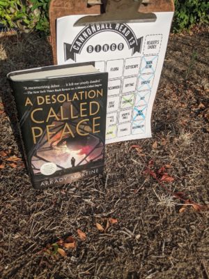 The book A Desolation Called Peace stands in front of a clipboard with a bingo sheet on it. The both are on dry ground with dead leaves and grasses.