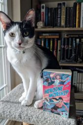 A black and white cat sitting next to the book Heroine Worship by Sarah Kuhn