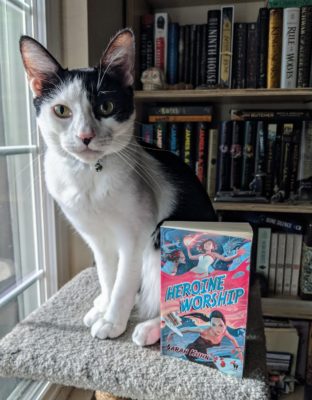A black and white cat sitting next to the book Heroine Worship by Sarah Kuhn