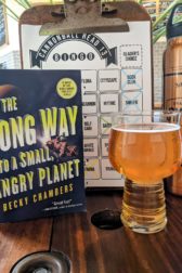 The book The Long Way to a Small, Angry Planet by Becky Chambers next to a pint glass of hard cider.