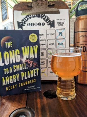 The book The Long Way to a Small, Angry Planet by Becky Chambers next to a pint glass of hard cider.