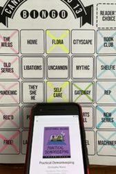 A phone with the audio book Practical Demonkeeping open on the screen, with a bingo board behind the phone.