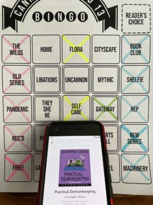 A phone with the audio book Practical Demonkeeping open on the screen, with a bingo board behind the phone.