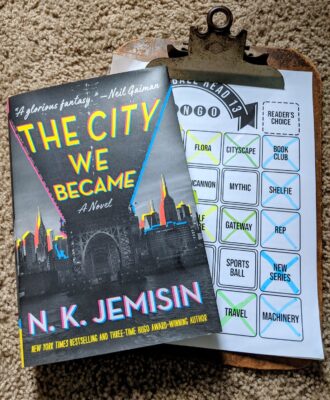 The book The City We Became by N.K. Jemisin lying on top of a clipboard holding a book bingo sheet