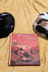 Two cats curled up, sleeping on a blanket with a book, "The Tea Dragon Tapestry" by K. O'Neill between them