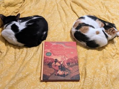 Two cats curled up, sleeping on a blanket with a book, "The Tea Dragon Tapestry" by K. O'Neill between them