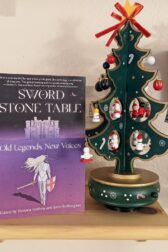 Picture of the book "Sword, Stone, Table: Old Legends, New Voice" is standing next to a miniature wooden Christmas tree hung with tiny ornaments.
