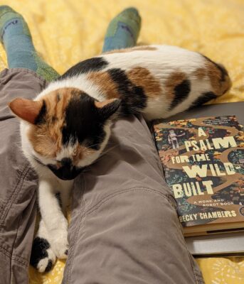 A calico cat sleeping on someone's stretched out legs with the book A Psalm for the Wild-Built by Becky Chambers.