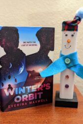 The book "Winter's Orbit" by Everina Maxwell stands next to a wooden snowman decoration.