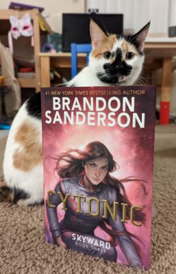 A calico cat sitting behind the book "Cytonic" by Brandon Sanderson.