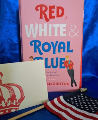 The book, "Red, White & Royal Blue" by Casey McQuiston standing behind a small American flag and a white notecard with an elaborate crown on it.