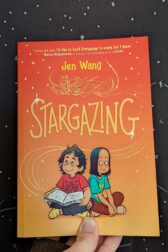 The book, "Stargazing" by Jen Wang held up in front of black fabric with little stars on it.