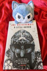 The book, "A Little Hatred" by Joe Abercrombie on a red satin background with a tiny, displeased looking plush wolf perched atop the book.