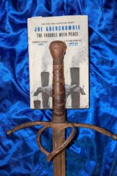 The book, "The Trouble with Peace" by Joe Abercrombie lying on a blue satin cloth with a rusty sword hilt lying on top of the book.
