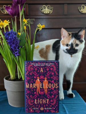 The book, "A Marvellous Light" by Freya Marske next to a pot of flowers. Behind them is a calico cat.