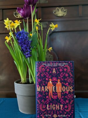 The book "A Marvellous Light" by Freya Marske standing next to a pot of daffodils, hyacinths, and tulips.
