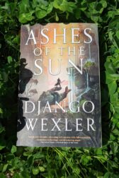The book, "Ashes of the Sun" by Django Wexler lying on top of a field of green clover.