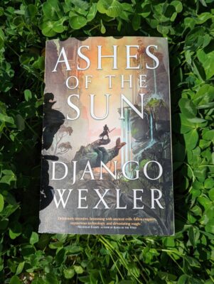 The book, "Ashes of the Sun" by Django Wexler lying on top of a field of green clover.