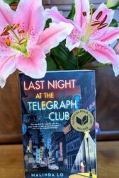 The book "Last Night at the Telegraph Club" by Malinda Lo in front of two blooming lilies.