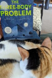 Calico cat curled up sleeping in front of the book, "The Three-Body Problem" by Cixin Liu.