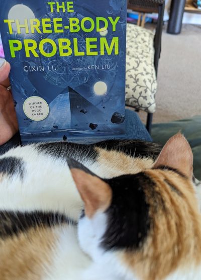 Calico cat curled up sleeping in front of the book, "The Three-Body Problem" by Cixin Liu.