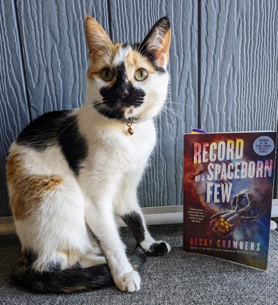 A calico cat sitting upright next to a the book, "Record of a Spaceborn Few" by Becky Chambers.