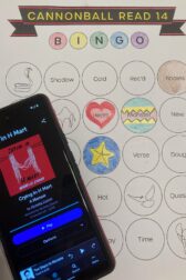 A cellphone showing audiobook, "Crying in H-Mart: A Memoir" by Michelle Zauner on the screen, lying on top of a book bingo card.