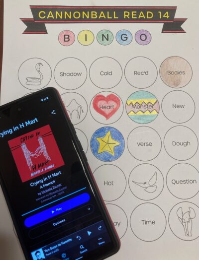 A cellphone showing audiobook, "Crying in H-Mart: A Memoir" by Michelle Zauner on the screen, lying on top of a book bingo card.