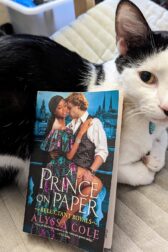 A black and white cat nestled on a grey chair. The book, "A Prince on Paper" by Alyssa Cole is propped up by resting on the cat's side.