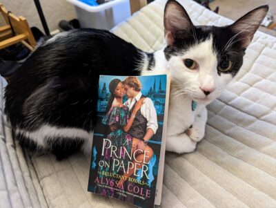 A black and white cat nestled on a grey chair. The book, "A Prince on Paper" by Alyssa Cole is propped up by resting on the cat's side.
