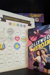 The book, "Hollywood Heroine" by Sarah Kuhn standing next to book bingo card.