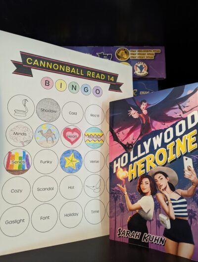 The book, "Hollywood Heroine" by Sarah Kuhn standing next to book bingo card.