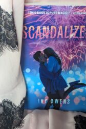 Pale pink fabric with black lace trim drapes around the book, "Scandalized" by Ivy Owens.