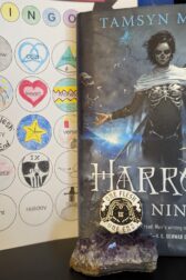 The book, "Harrow the Ninth" by Tamsyn Muir standing next to a book bingo card. In front of the book is a pin resting on a small bed of amethyst. The pin says, "One Flesh, One End" with skeletal hands in the shape of a heart with the roman numerals for ten in the center.