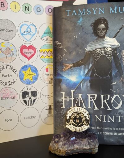 The book, "Harrow the Ninth" by Tamsyn Muir standing next to a book bingo card. In front of the book is a pin resting on a small bed of amethyst. The pin says, "One Flesh, One End" with skeletal hands in the shape of a heart with the roman numerals for ten in the center.
