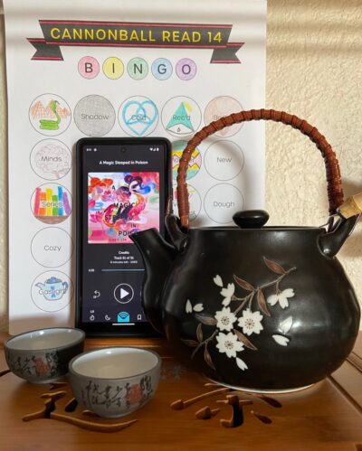 A small black tea pot with white flowers on it sits next to two tiny dark brown tea cups. Behind them is a cell phone showing the image of an audiobook. The audio book is "A Magic Steeped in Poison" by Judy I. Lin. In the background is a book bingo card.