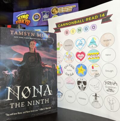 The book, "Nona the Ninth" by Tamsyn Muir standing next to a mostly filled out book bingo card.