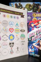 The book "Holiday Heroine" by Sarah Kuhn standing next to a mostly full book bingo card.