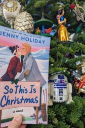 The book, "So This is Christmas" by Jenny Holiday being held up in front of a Christmas tree. Next to the book is an ornament of the robot R2D2 from Star Wars.
