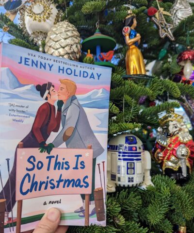 The book, "So This is Christmas" by Jenny Holiday being held up in front of a Christmas tree.  Next to the book is an ornament of the robot R2D2 from Star Wars.