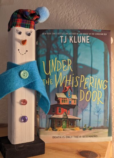 The book, "Under the Whispering Door" by TJ Klune standing next to a wooden snowman.