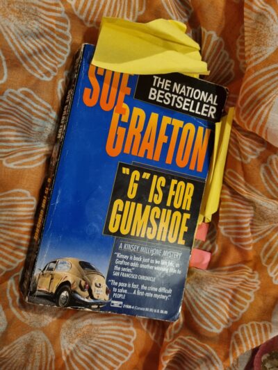 Paperback of G is for Gumshoe on an orange scarf, blue cover showing a VW car, with orange text