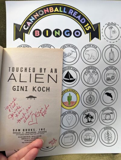 A hand holding open the book, "Touched by an Alien" by Gini Koch on the title page with a message written in hand. "Hannah and Ted, join the team, touch an alien. Gini Koch". The book is in front of a book bingo card with the North America circle colored.
