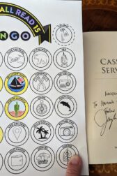 The book, "Cassiel's Servant" open to the title page. There is a handwritten note, "To Hannah and Ted, follow your hearts!", signed by Jacqueline Carey. This is next to a partially filled out book bingo card.