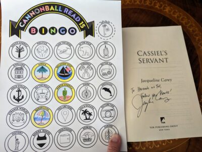 The book, "Cassiel's Servant" open to the title page. There is a handwritten note, "To Hannah and Ted, follow your hearts!", signed by Jacqueline Carey. This is next to a partially filled out book bingo card.